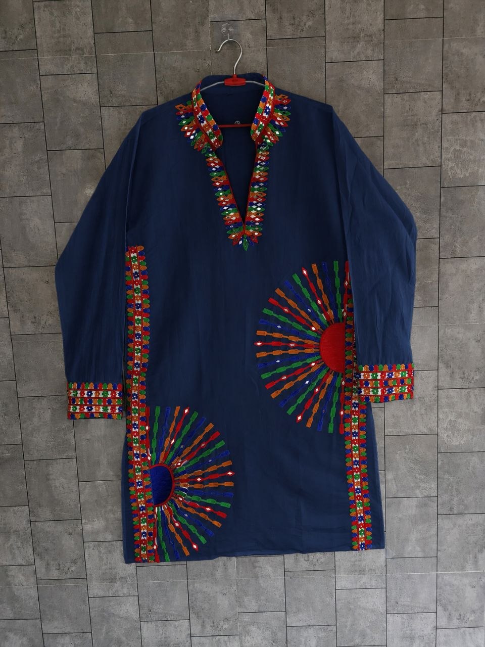 A navy blue long-sleeve tunic or kurta with colorful geometric embroidered patterns around the collar, cuffs, hem, and large sunburst motifs on the front, hanging against a gray tiled wall.