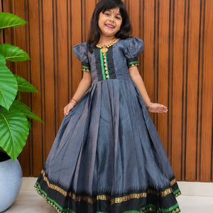 A young girl modeling a dark gray-blue traditional Indian dress with green and gold embroidery, standing in front of a brown wooden slatted wall.