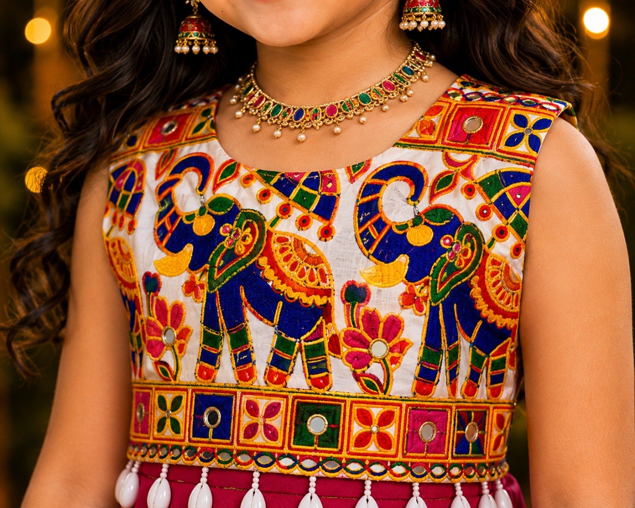 A close-up of a child wearing a sleeveless traditional outfit with colorful elephant and floral embroidery, along with matching earrings and a necklace.