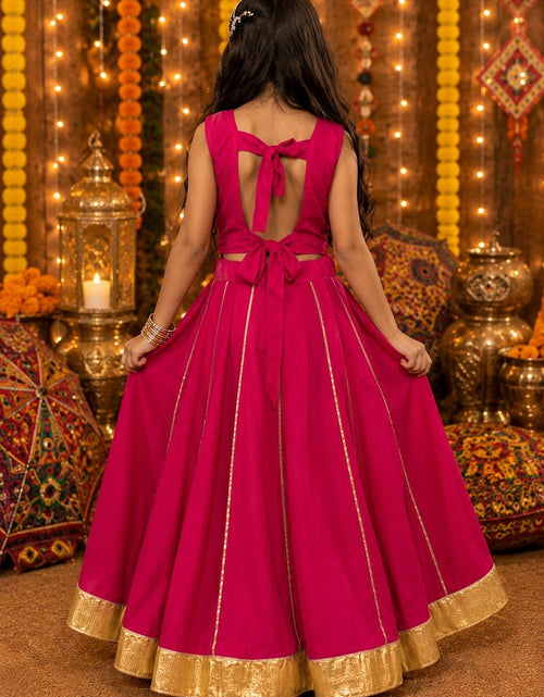 Load image into Gallery viewer, A young girl viewed from behind wearing a bright magenta pink traditional Indian gown with gold zari borders and a bow detail on the back, standing in a decorated festive setting with warm lights and marigold garlands.

