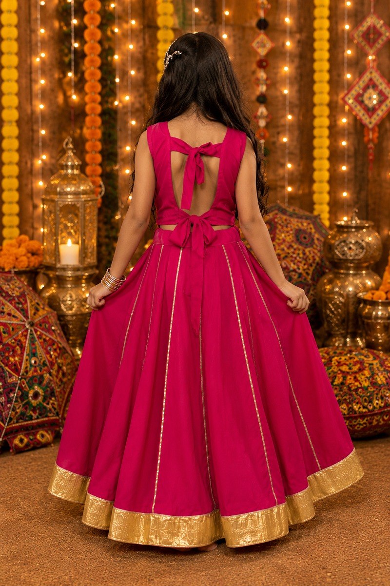 A young girl viewed from behind wearing a bright magenta pink traditional Indian gown with gold zari borders and a bow detail on the back, standing in a decorated festive setting with warm lights and marigold garlands.