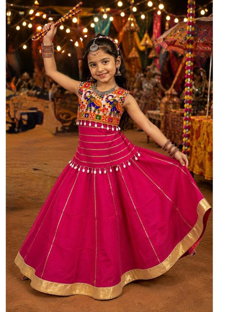 Load image into Gallery viewer, A young girl wearing a bright magenta pink traditional Indian lehenga choli dress with gold borders and colorful embroidered details on the bodice, posing and twirling in a festive indoor setting with string lights and decorations.
