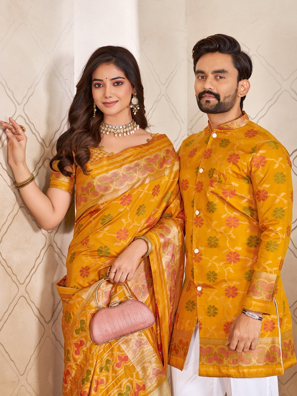 A couple wearing traditional Indian formal wear, featuring a woman in a yellow and gold patterned saree and a man in a matching yellow embroidered kurta, standing against a beige patterned wall.