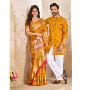 A man and a woman posing in traditional Indian attire. The woman wears a yellow and gold Banarasi silk saree with pink and orange floral patterns and a pink clutch.