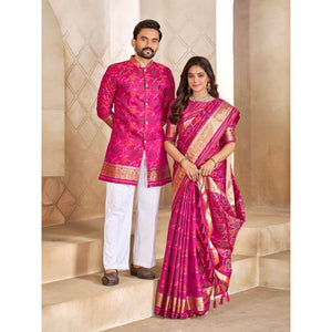 A man and a woman modeling traditional Indian formal wear. The man wears a bright pink embroidered kurta with white trousers, and the woman wears a matching pink and gold embroidered saree, standing on beige stone steps.