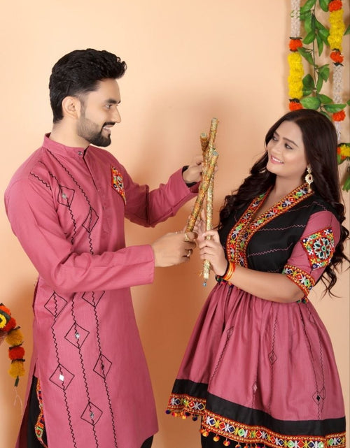 Load image into Gallery viewer, A man and a woman wearing traditional Indian Navratri ethnic wear, specifically pink and black kurta sets with colorful embroidery, holding a decorative wooden object against a peach-colored wall decorated with floral garlands.
