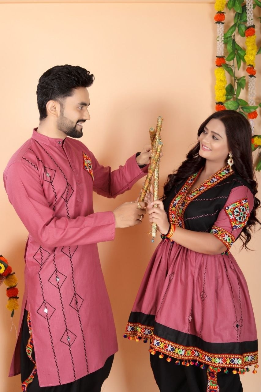 A man and a woman wearing traditional Indian Navratri ethnic wear, specifically pink and black kurta sets with colorful embroidery, holding a decorative wooden object against a peach-colored wall decorated with floral garlands.