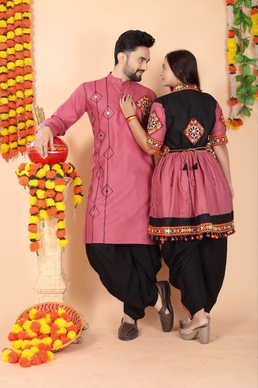 A man and a woman wearing matching traditional Indian ethnic wear, featuring pink and black kurta sets with colorful embroidery, posed in front of a decorated peach-colored backdrop with marigold garlands.