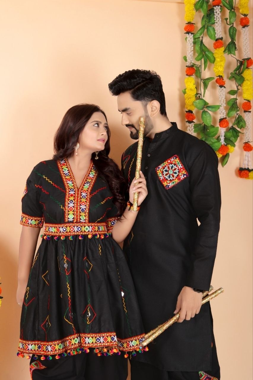 A couple posing in traditional Indian ethnic Navratri wear. The woman wears a black kurta with colorful geometric embroidery, and the man wears a matching black kurta with a similar embroidered patch on the chest.