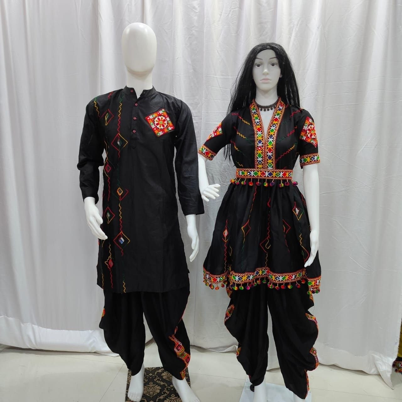 A traditional black embroidered couple wear Navratri kurta and pant set displayed on two mannequins against a white curtain backdrop. The Matching set features vibrant red, yellow, and green geometric embroidery on the chest, sleeves, and hem. 