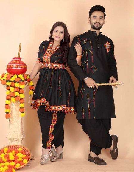 A man and a woman modeling a matching black traditional Indian Navratri outfit set with red and gold embroidered details, standing in front of a beige studio backdrop next to a decorative pot and flower garlands.