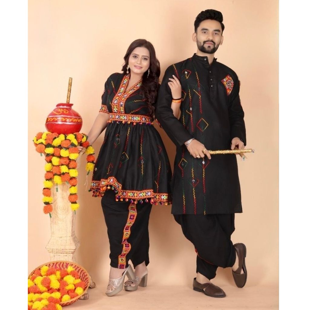 A man and a woman modeling a matching black traditional Indian Navratri outfit set with red and gold embroidered details, standing in front of a beige studio backdrop next to a decorative pot and flower garlands.