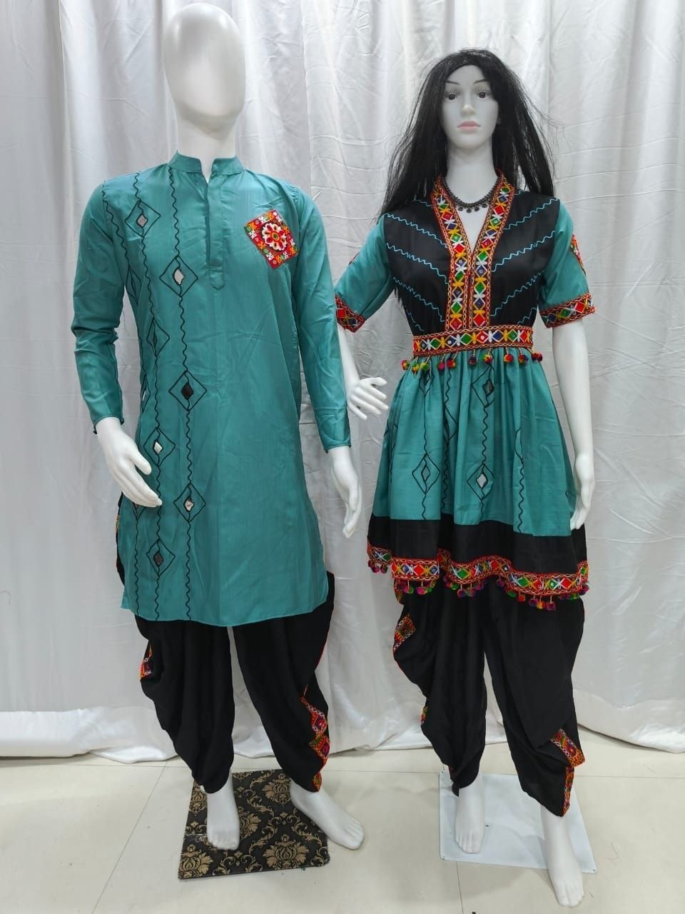 A traditional Indian Couple wear matching outfit displayed on two mannequins, featuring a teal long kurta with black harem pants on the left, and a matching teal and black tunic with a flared skirt and embroidered borders on the right.