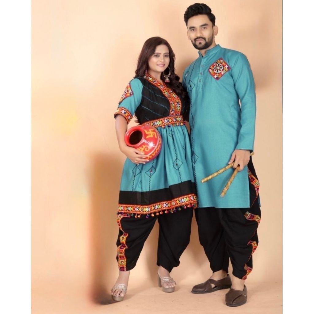 A couple modeling traditional Navratri Matching Outfit clothing against a solid beige studio backdrop. The woman wears a turquoise and black kurta with colorful embroidered borders and black pants, holding a red pot.