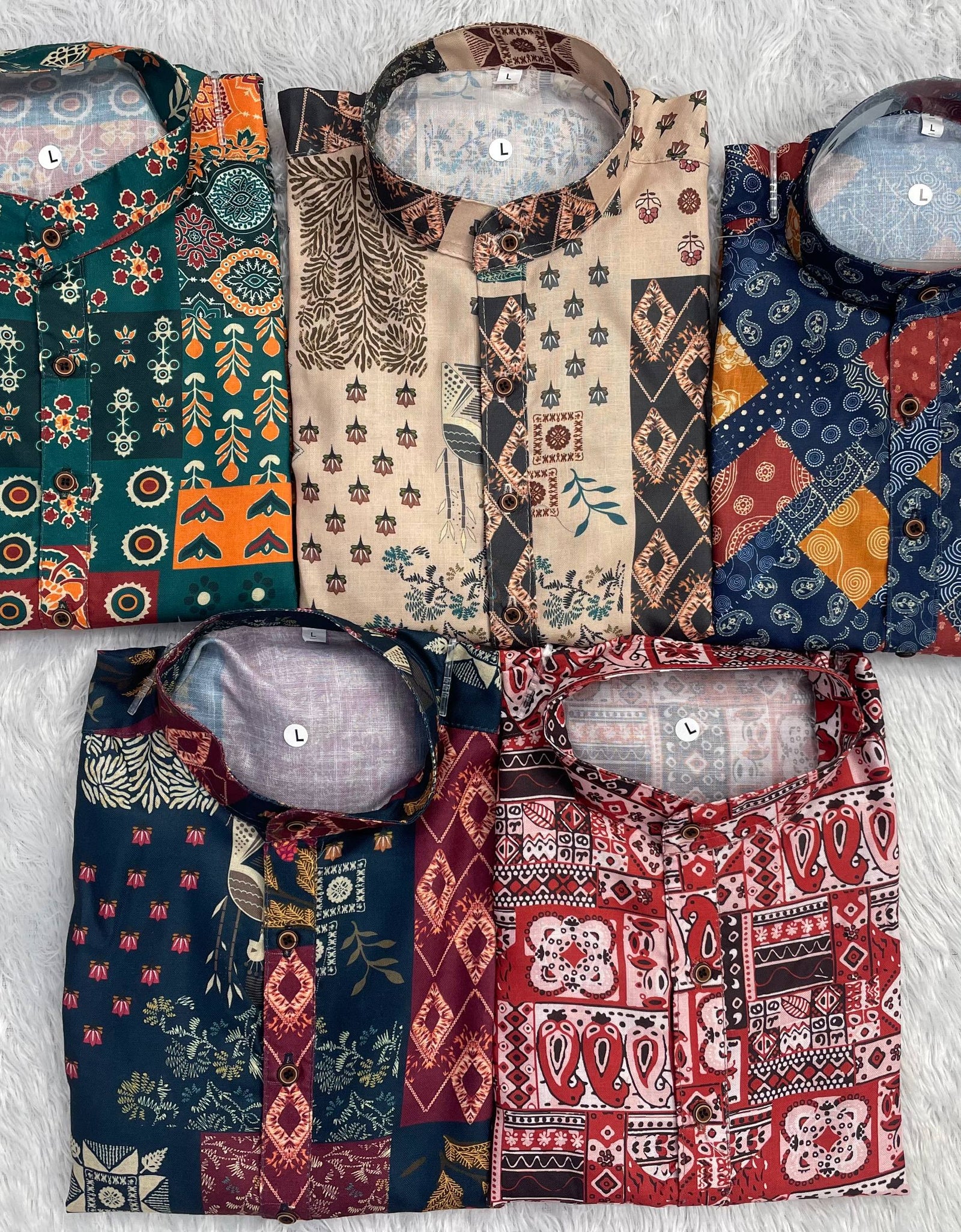 Five folded patterned kurta shirts arranged in a 2x3 grid on a white faux-fur surface. Each shirt has a different multicolor patchwork print with geometric, floral, and tribal motifs in orange, teal, beige, navy, and red.