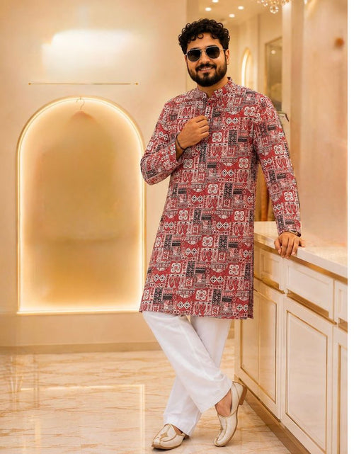 Load image into Gallery viewer, A man wearing a red and black patterned kurta with white trousers and gold shoes, standing in a modern, warm-toned interior hallway.
