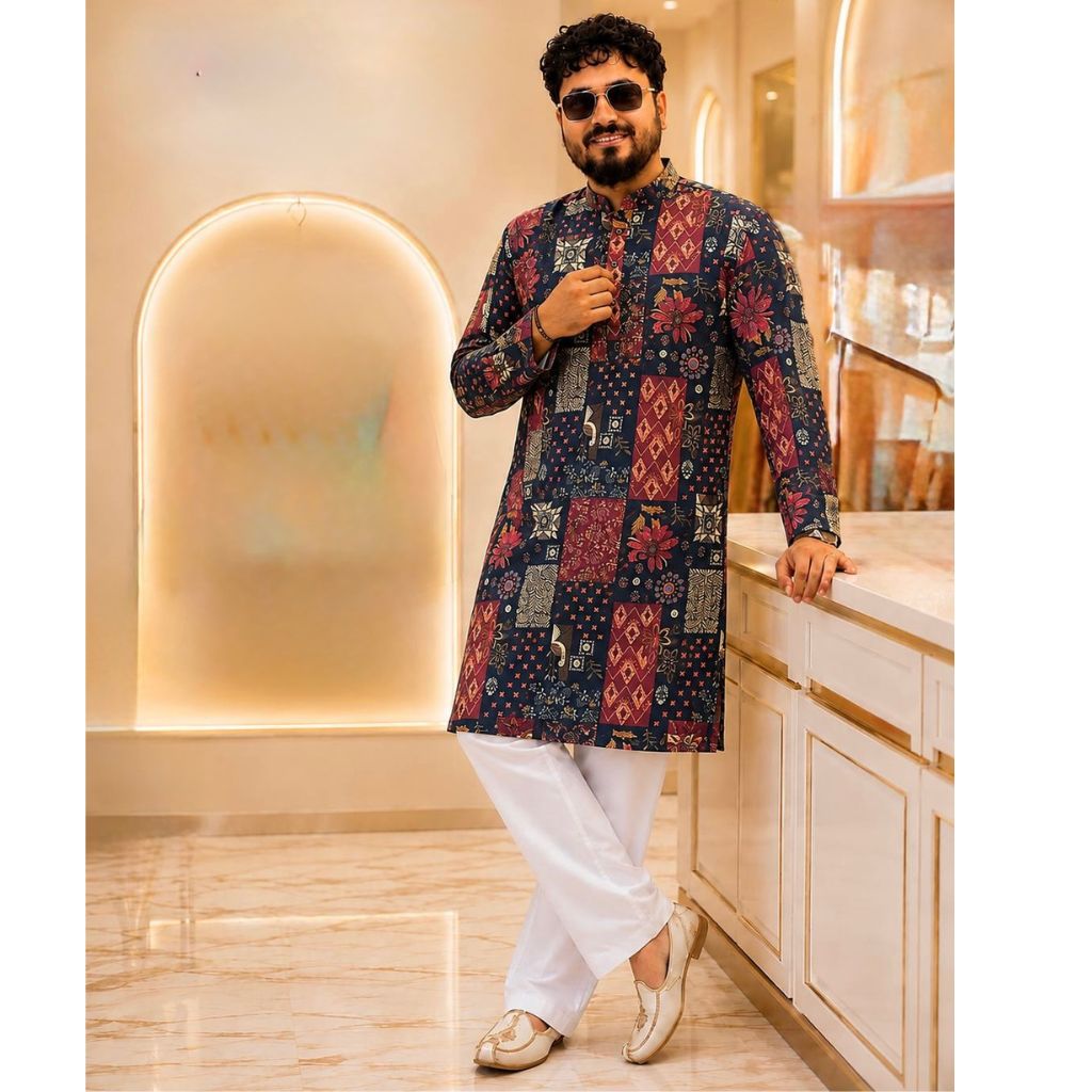 A man wearing a long-sleeved kurta with red and gold patchwork patterns, paired with white trousers and gold shoes, posing in a modern, warm-toned interior setting.