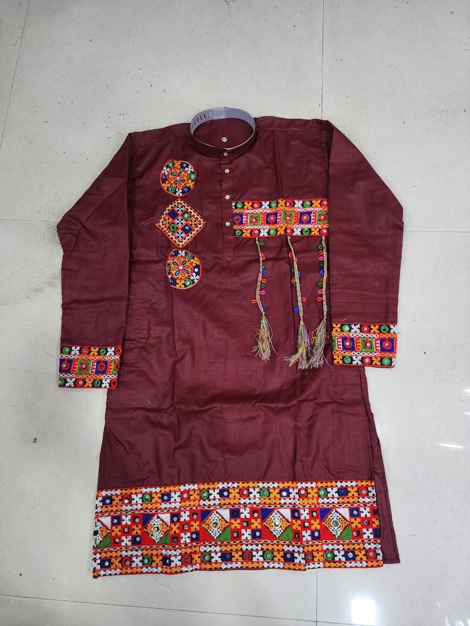 A maroon long-sleeve kurta with colorful geometric embroidery and tassels, laid flat on a light-colored tiled floor.