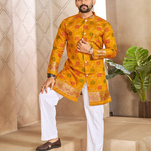 A man modeling a bright yellow-orange kurta with intricate multicolor floral patterns and white trousers, standing on beige stone steps in front of a patterned beige wall.