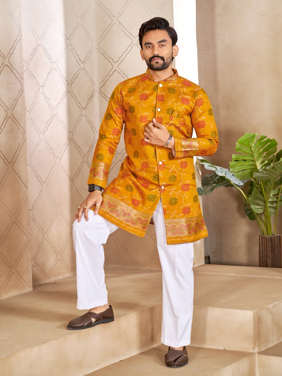 A man modeling a bright yellow-orange kurta with intricate multicolor floral patterns and white trousers, standing on beige stone steps in front of a patterned beige wall.