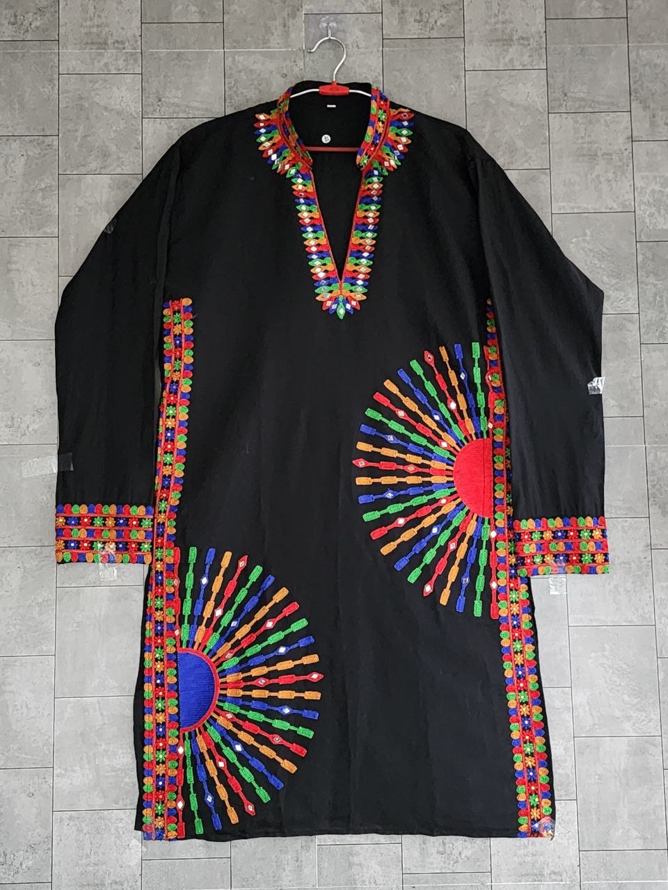 A black long-sleeve tunic or kaftan with vibrant multicolor embroidered geometric patterns, shown hanging against a gray tiled wall.
