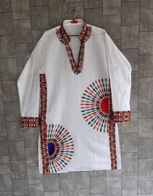 Load image into Gallery viewer, A white long-sleeve tunic or kaftan hanging on a hanger against a gray tiled wall. The garment features colorful geometric embroidered patterns around the neckline, cuffs, hem, and two large sunburst motifs on the front.
