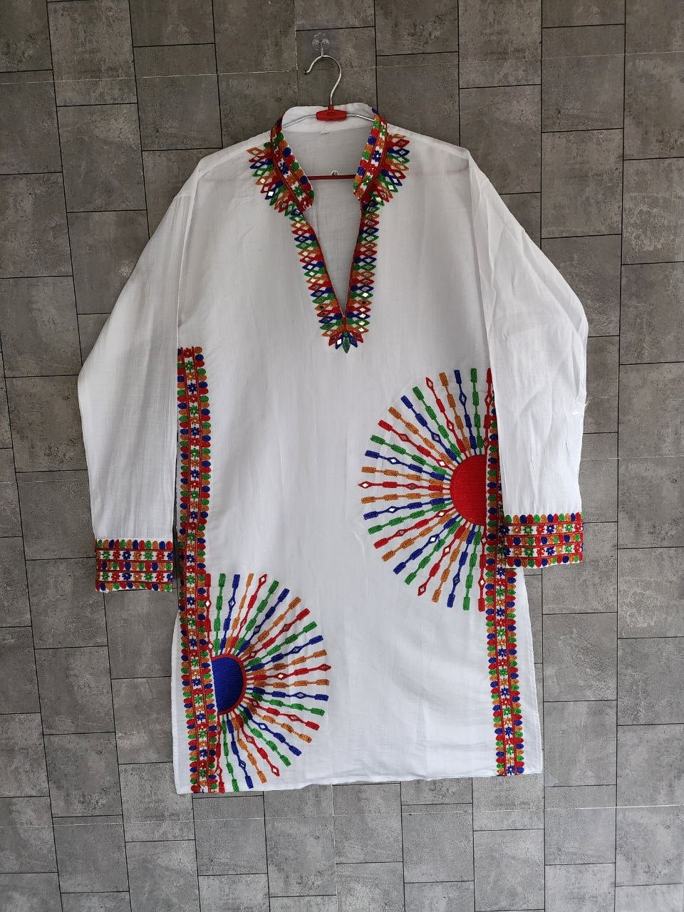 A white long-sleeve tunic or kaftan hanging on a hanger against a gray tiled wall. The garment features colorful geometric embroidered patterns around the neckline, cuffs, hem, and two large sunburst motifs on the front.