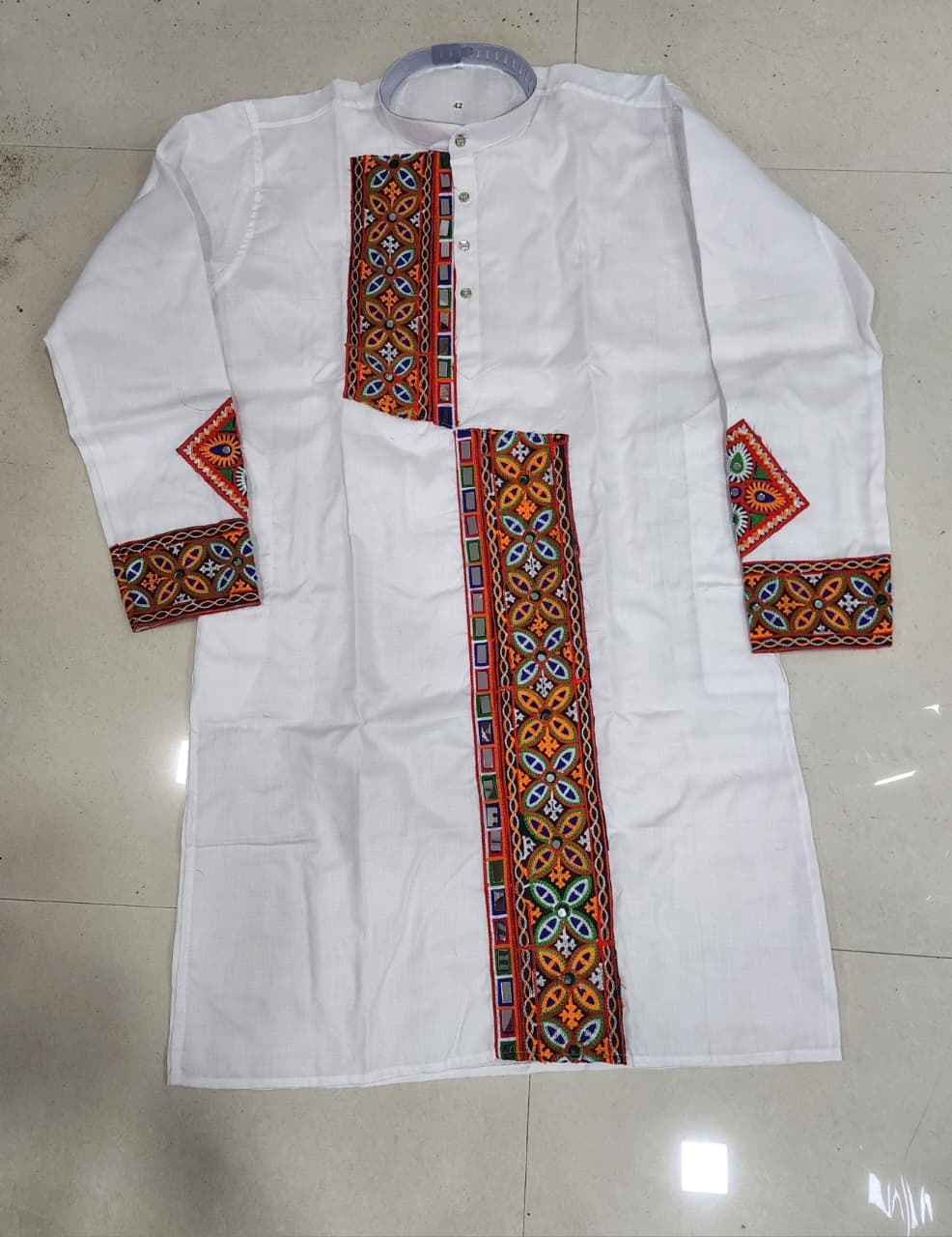 A white long-sleeve kurta laid flat on a light beige tiled floor, featuring colorful geometric embroidered patterns in red, orange, blue, and green along the center placket, cuffs, and collar.