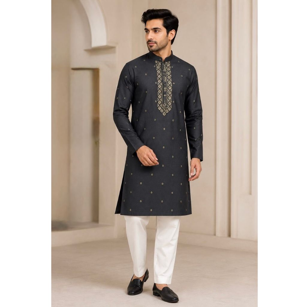 A man modeling a dark charcoal gray long-sleeve kurta with gold geometric embroidery on the chest, paired with white trousers and black loafers, set against a beige architectural background.