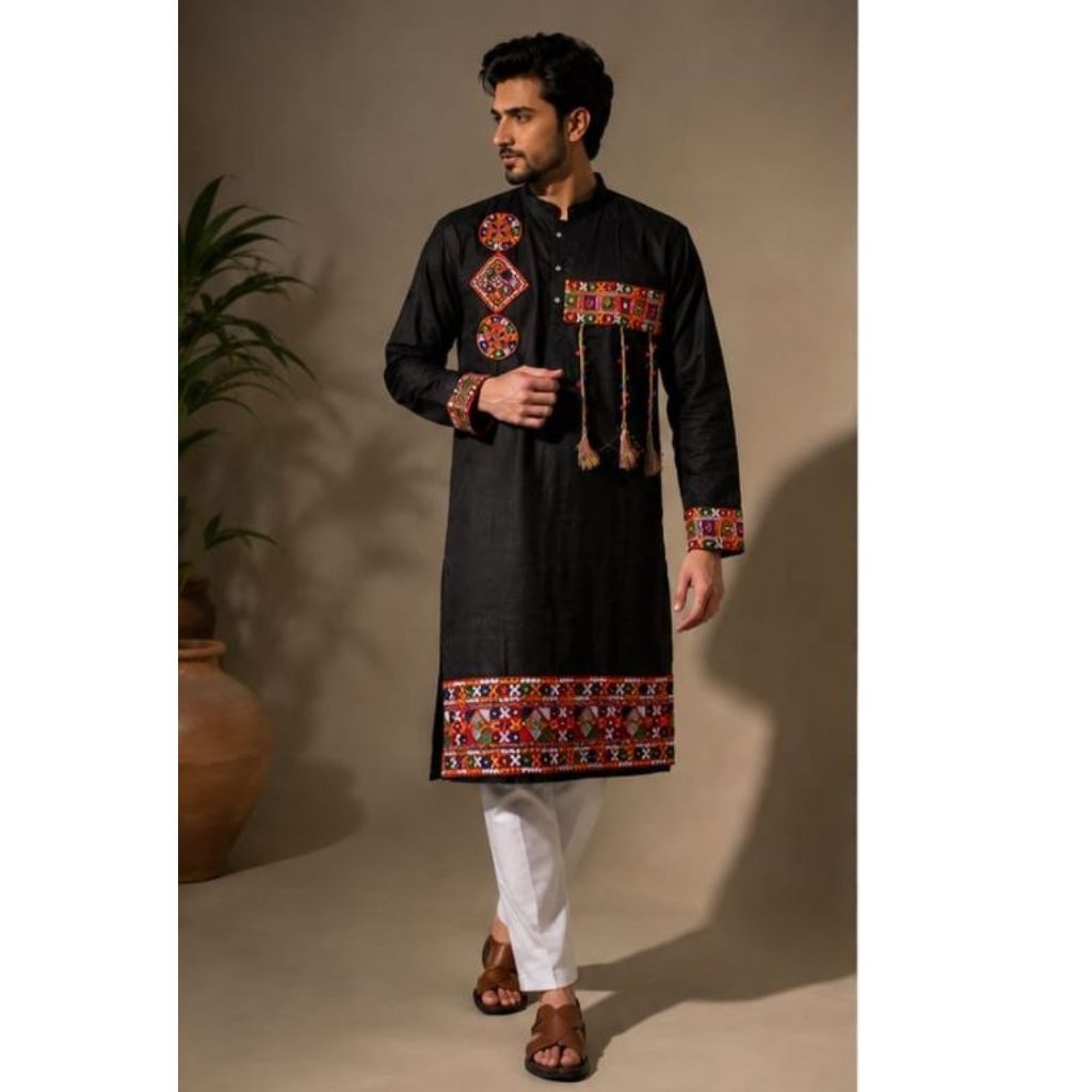 A man modeling a Navratri Festive wear black long kurta with colorful embroidered patterns on the chest, cuffs, and hem, paired with white trousers and brown sandals in a studio setting.