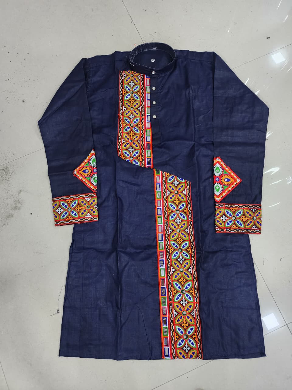 A navy blue long-sleeve kurta with colorful geometric embroidered patterns on the front placket, cuffs, and collar, laid flat on a light gray tiled floor.