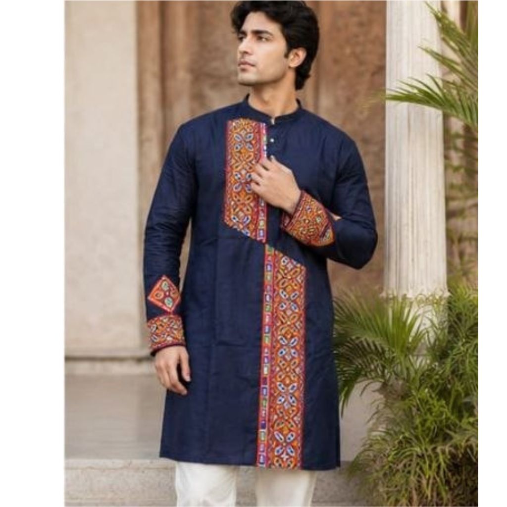 A man wearing a navy blue kurta with colorful geometric embroidered panels on the front and cuffs, paired with white trousers, standing outdoors in front of a stone column and plants.