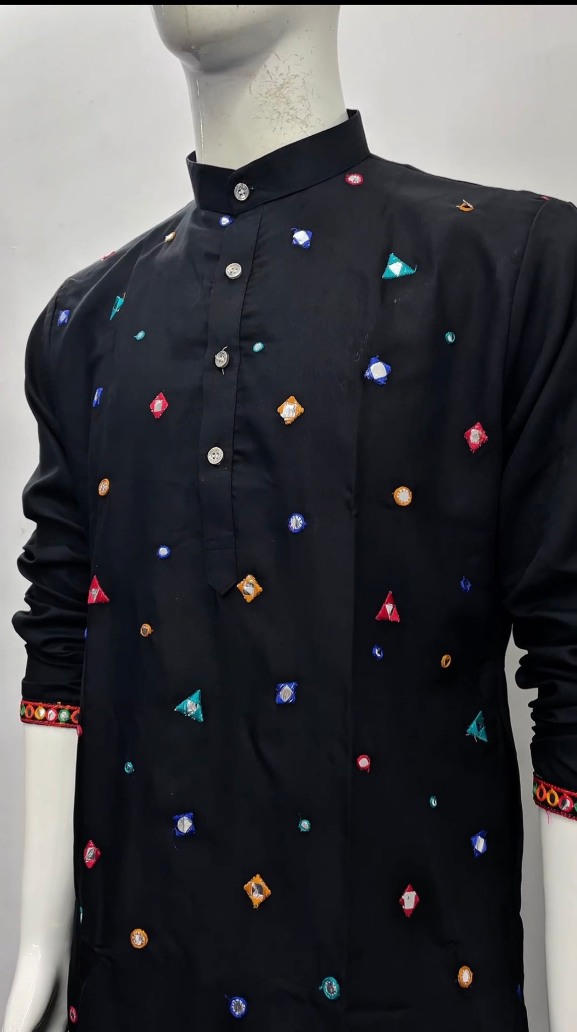 A black long-sleeve kurta displayed on a white mannequin, featuring colorful geometric embroidered motifs in red, blue, orange, and green, with a mandarin collar and buttoned front.