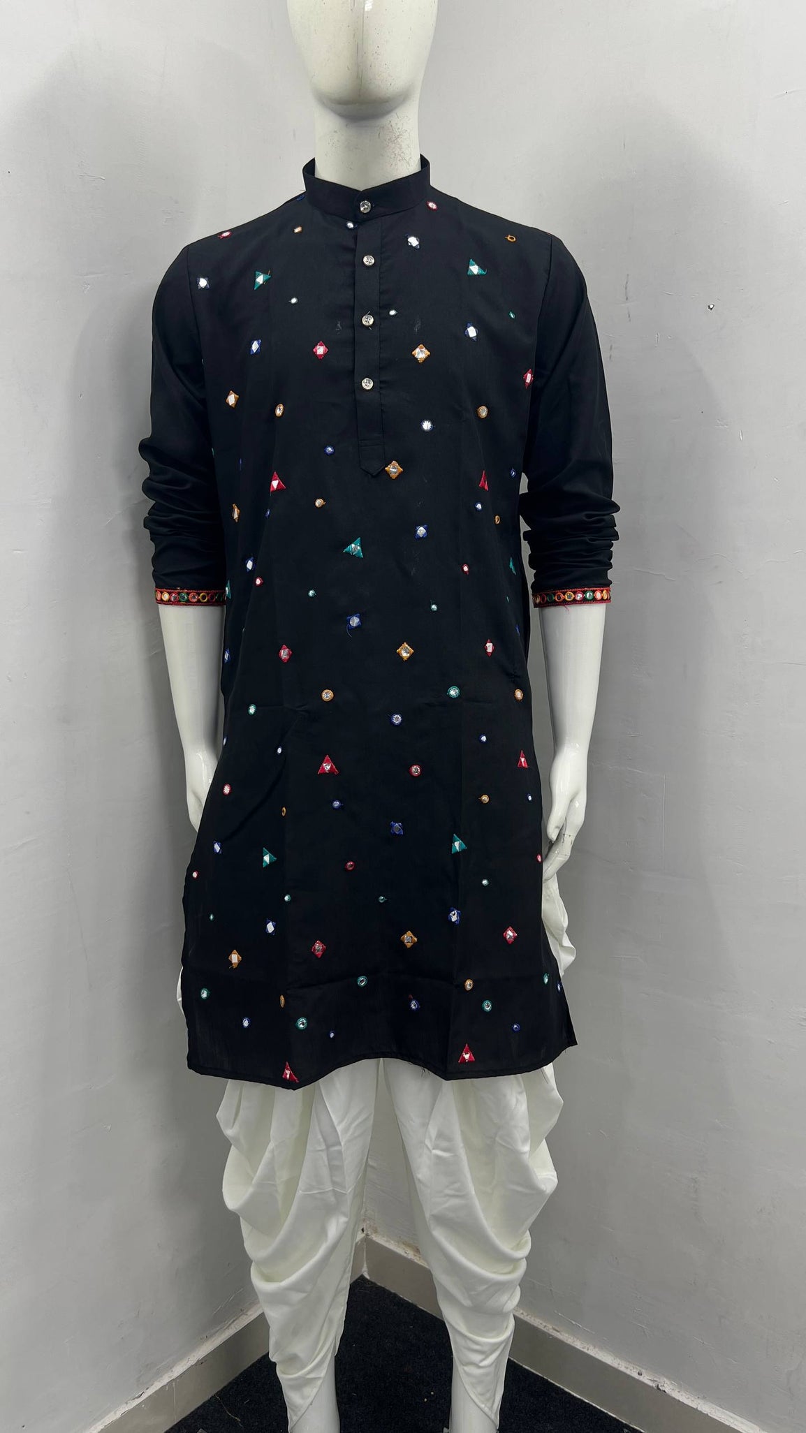 A black long-sleeve kurta with colorful embroidered geometric patterns, displayed on a white mannequin with white dhoti pants against a plain gray wall.
