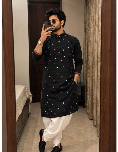 A man takes a mirror selfie wearing a black long-sleeve kurta decorated with small colorful embroidered motifs, paired with white pants and black shoes, in an indoor room setting.