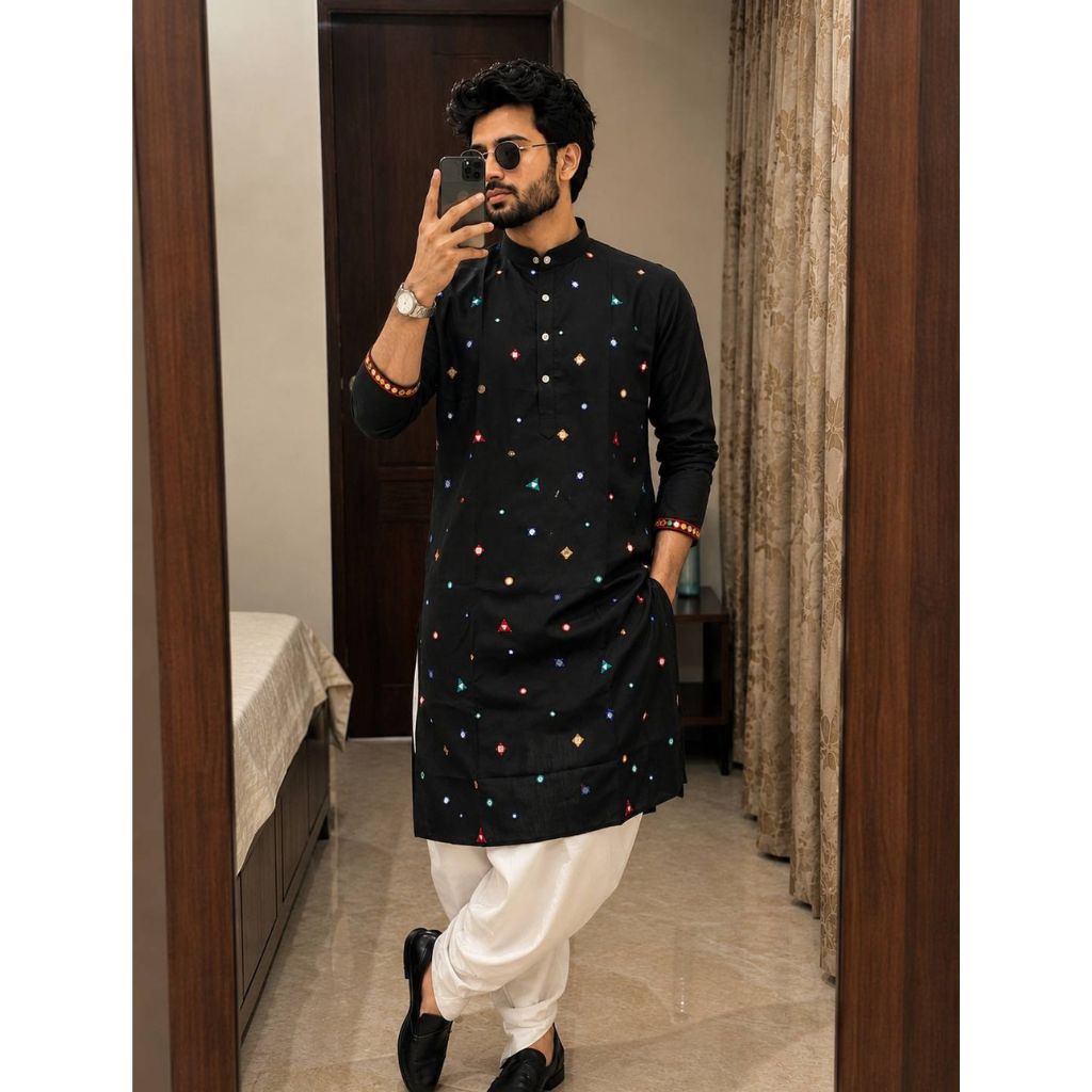 A man takes a mirror selfie wearing a black long-sleeve kurta decorated with small colorful embroidered motifs, paired with white pants and black shoes, in an indoor room setting.