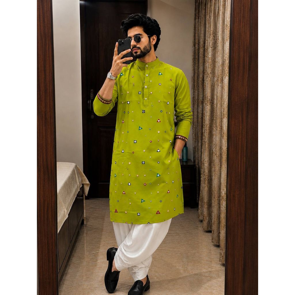A man takes a mirror selfie wearing a bright lime green kurta with small colorful geometric prints, paired with white trousers and black shoes in an indoor setting.