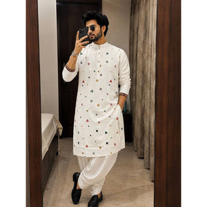 A man takes a mirror selfie wearing a white long-sleeve Indian traditional kurta with small colorful geometric prints, paired with white trousers and black shoes, in an indoor room setting.