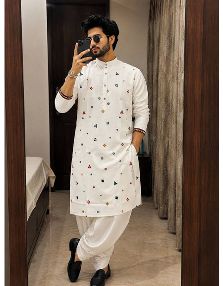 A man takes a mirror selfie wearing a white long-sleeve Indian traditional kurta with small colorful geometric prints, paired with white trousers and black shoes, in an indoor room setting.