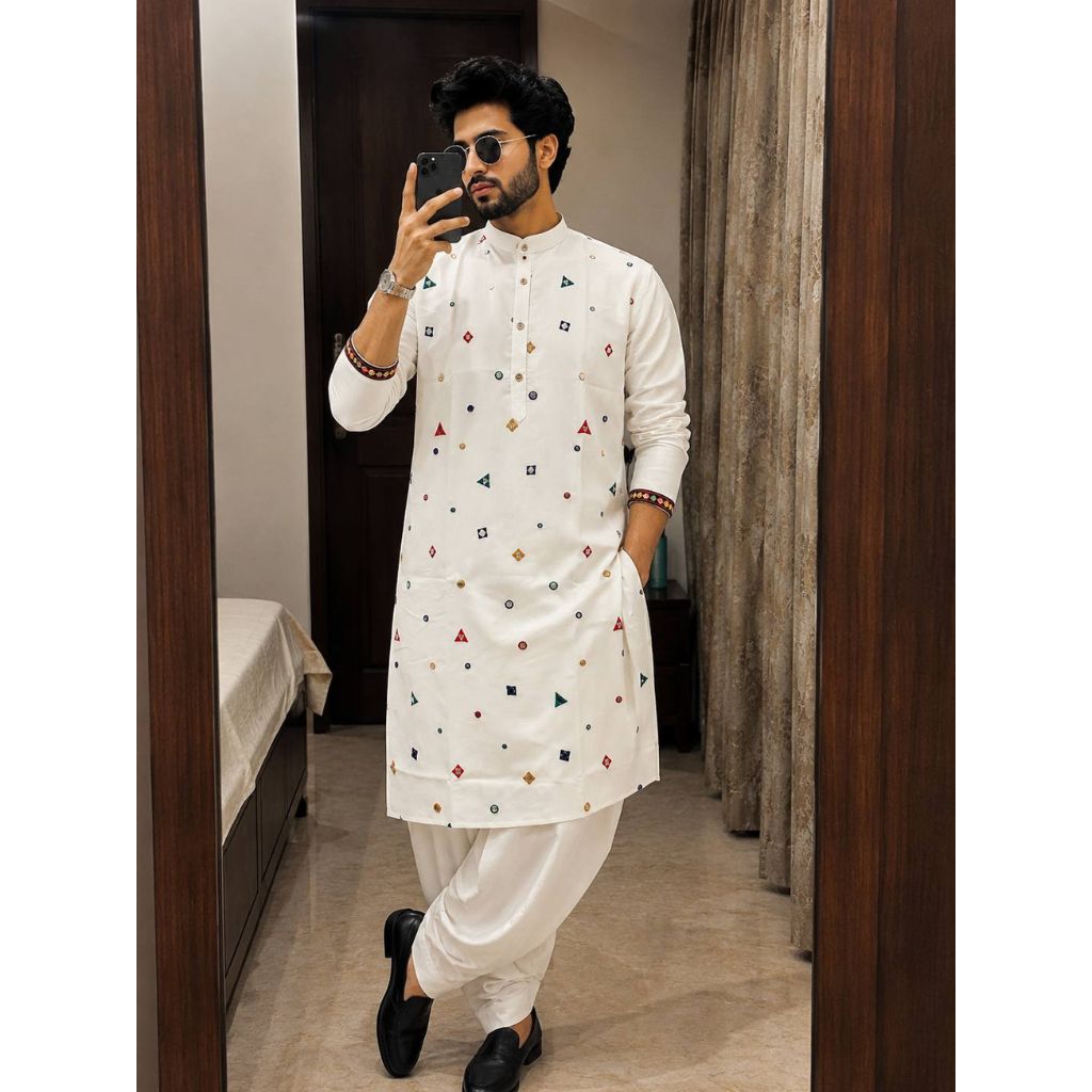 A man takes a mirror selfie wearing a white long-sleeve Indian traditional kurta with small colorful geometric prints, paired with white trousers and black shoes, in an indoor room setting.