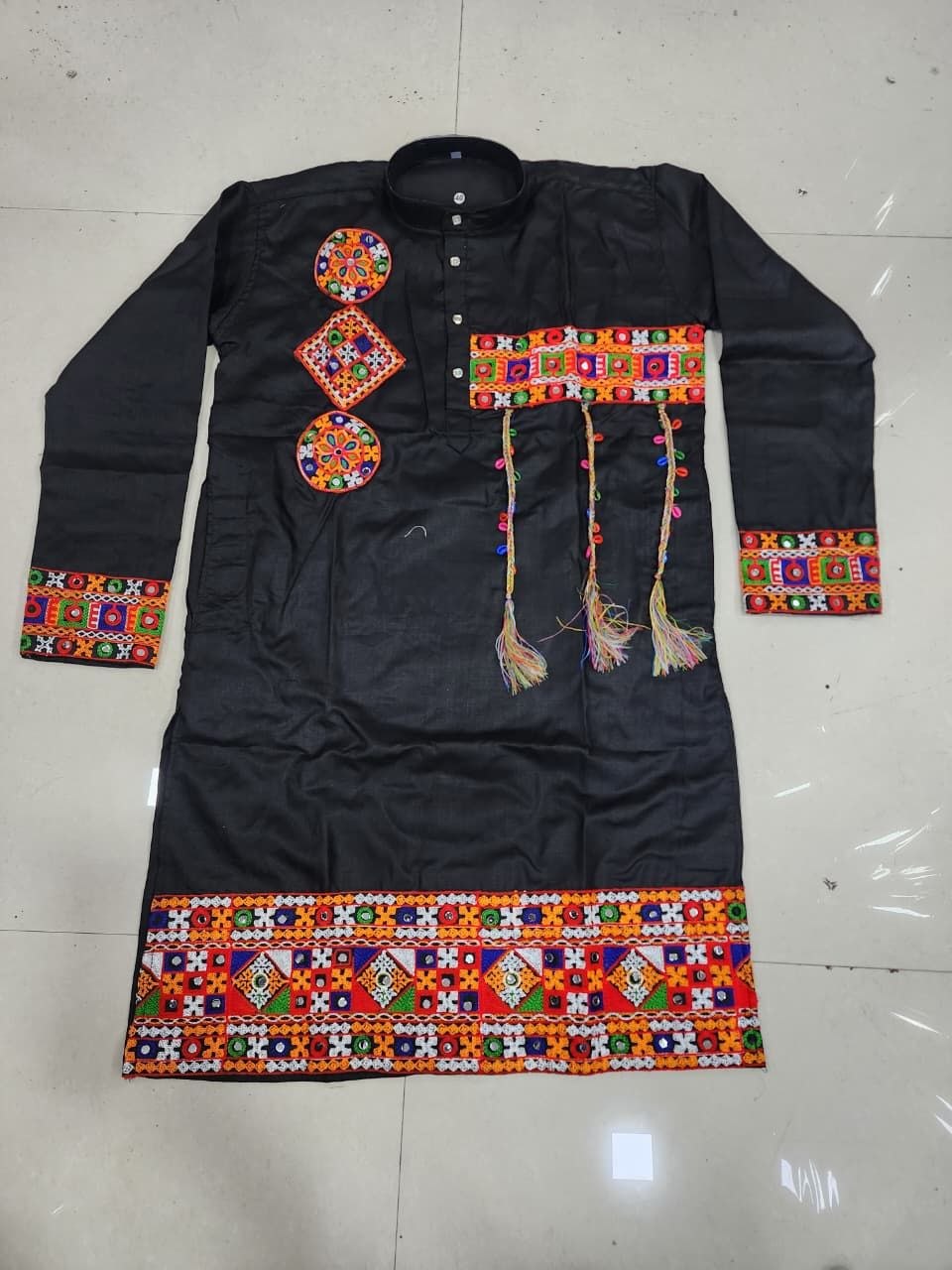 A black long-sleeve traditional tunic or kurta laid flat on a light tiled floor, decorated with colorful geometric embroidered patterns on the chest, cuffs, hem, and hanging tassels.