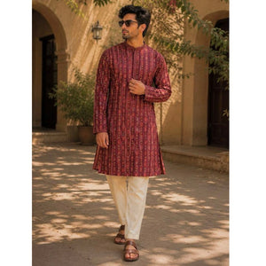 A man wearing a festive wear ethnic traditional maroon patterned long-sleeve kurta and cream-colored pants walks outdoors in front of a beige stone building with arches and trees casting shadows.