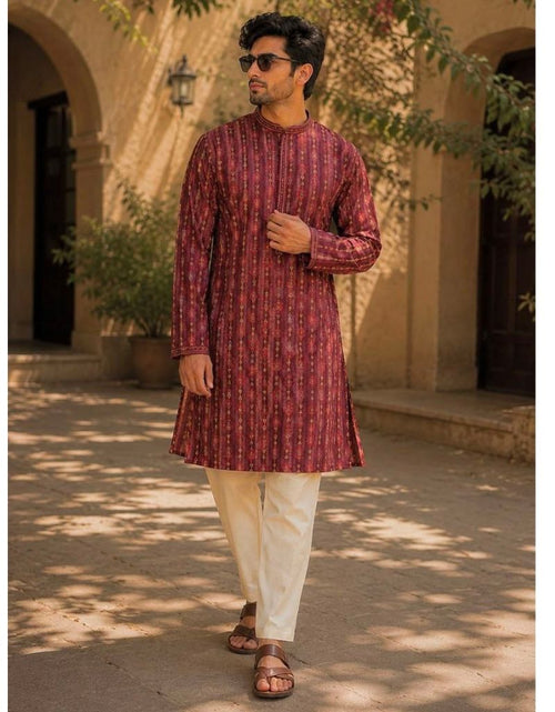 Load image into Gallery viewer, A man wearing a festive wear ethnic traditional maroon patterned long-sleeve kurta and cream-colored pants walks outdoors in front of a beige stone building with arches and trees casting shadows.

