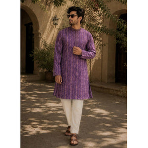 A man wearing a festive wear ethnic traditional Indian purple patterned kurta with white trousers and brown sandals, standing outdoors in front of a beige stone building with arches and leafy branches.