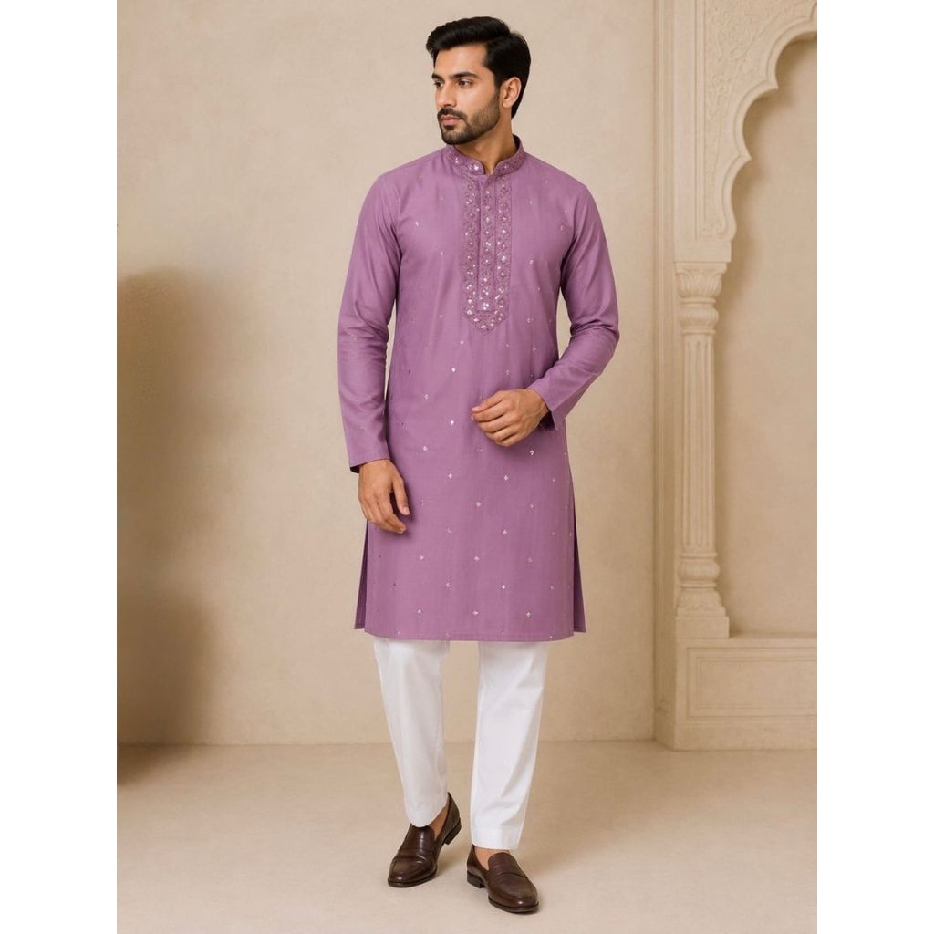 A man wearing a lavender purple kurta with silver embroidery on the chest and white trousers, standing in front of a beige architectural background.