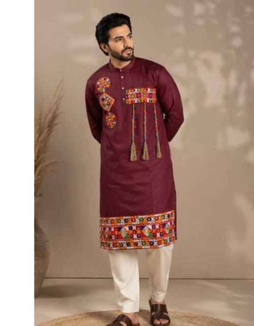Load image into Gallery viewer, A man modeling a festive wear Navratri deep maroon kurta with colorful geometric embroidery on the chest and hem, paired with white trousers and brown sandals, set against a warm beige background with soft leaf shadows.
