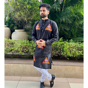 A man wearing a black kurta with colorful geometric embroidered patterns on the chest and lower hem, paired with white trousers and black shoes, standing outdoors in front of green plants and potted plants.