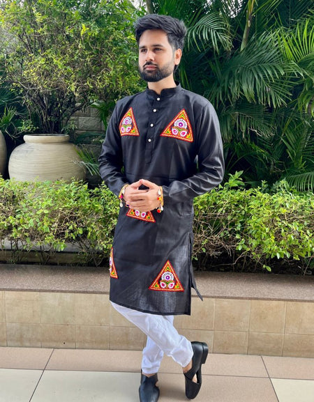 A man wearing a black kurta with colorful geometric embroidered patterns on the chest and lower hem, paired with white trousers and black shoes, standing outdoors in front of green plants and potted plants.