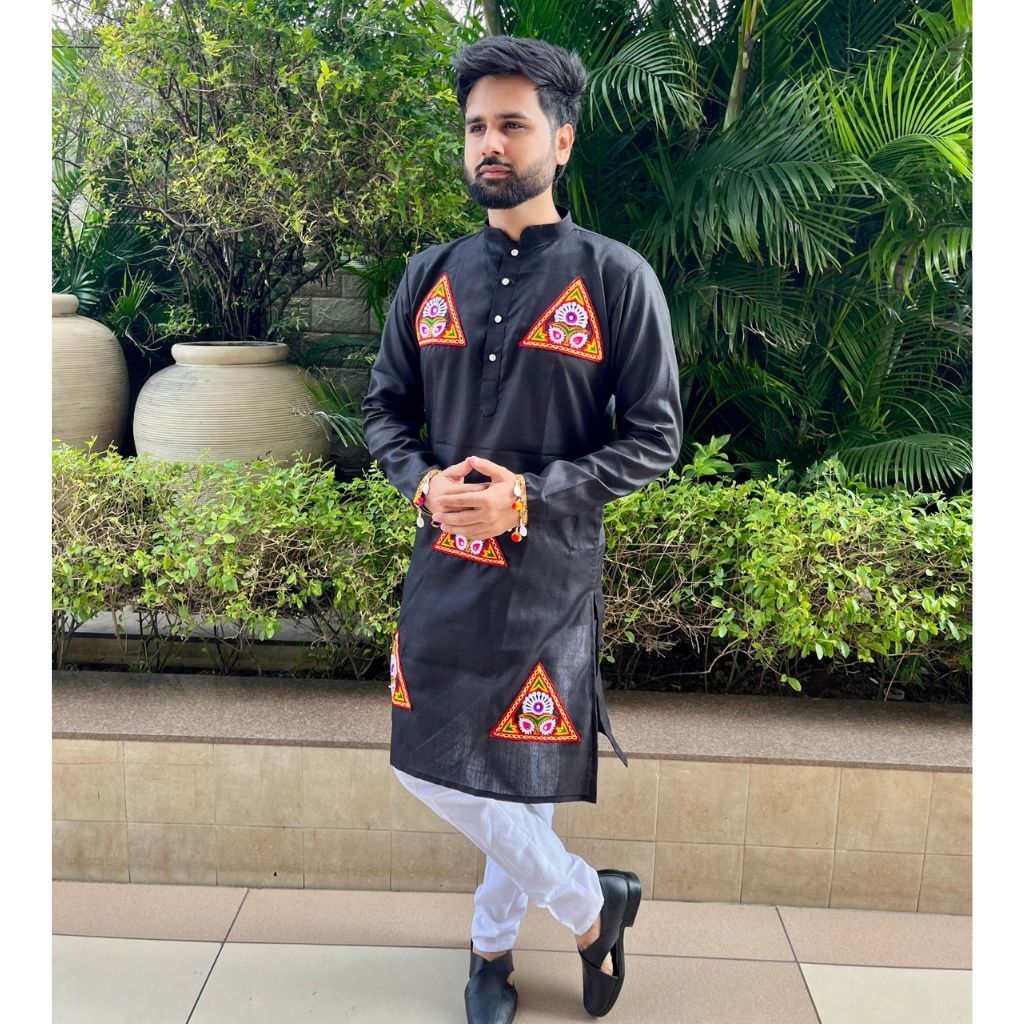 A man wearing a black kurta with colorful geometric embroidered patterns on the chest and lower hem, paired with white trousers and black shoes, standing outdoors in front of green plants and potted plants.