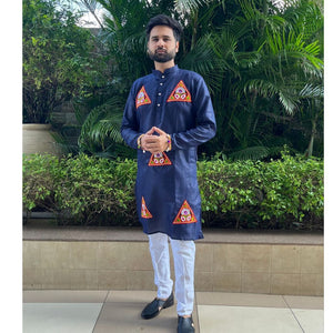 A man wearing a navy blue kurta with red and gold geometric patterns on the chest and lower hem, paired with white trousers and black shoes, standing outdoors in front of green plants.